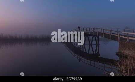 Impressionen einer Frühlingswanderung bei Sonnenaufgang und Nebel in Südholland in der Region Alblasserwaard Vijfheerenlanden bei Kinderdijk: Eine Person geht über eine Fußgängerbrücke Stockfoto
