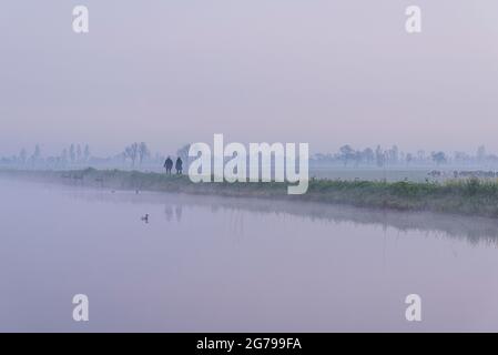 Eindrücke einer Frühlingswanderung bei Sonnenaufgang und Nebel in Südholland in der Region Alblasserwaard Vijfheerenlanden bei Kinderdijk: Zwei Wanderer entlang eines Kanals, Silhouetten Stockfoto