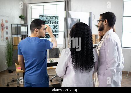 Vier multiethnische Ärzte analysieren einen Scan oder Röntgenfilm oder erklären einen CT-Scan zusammen mit ernsthaften, durchdachten Äußerungen. Diagnose, Behandlungsergebnisse, Anomalie auf dem Film Stockfoto