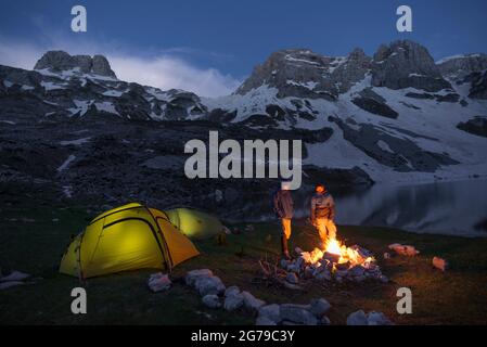 Zwei Wanderer stehen am Lagerfeuer neben ihren beleuchteten Zelten im Prokletije Nationalpark in der Nähe eines Bergsees in Albanien Stockfoto