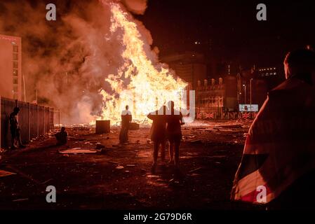 Belfast, Großbritannien. Juli 2021. Zuschauer beobachten das Lagerfeuer bei den Feierlichkeiten zur elften Nacht in Sandy Row, Belfast. Um die Schlacht von Boyne zu feiern, werden in verschiedenen protestantischen Vierteln in Belfast Lagerfeuer errichtet und Straßen und Häuser mit Union Jacks geschmückt. Die Lagerfeuer werden in der Nacht vor den Paraden des 12. Juli, am 11. Juli, angezündet. (Foto von Natalia Campos/SOPA Images/Sipa USA) Quelle: SIPA USA/Alamy Live News Stockfoto