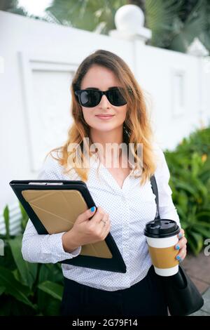 Porträt einer positiven Geschäftsfrau in Sonnenbrillen, die Kaffeebecher und Dokumentenmappe hält, wenn sie im Freien steht Stockfoto