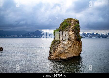 Der Blick von Ponte da Boa Viagem, Nitreói, Bundesstaat Rio de Janeiro, Brasilien. Grüne Ilha da Boa Viagem Privatinsel in Bucht, die über eine schmale Brücke und gelben Sand unter blauer Wolkenlandschaft mit dem Hauptland verbunden ist. Berge in der Ferne. Stockfoto