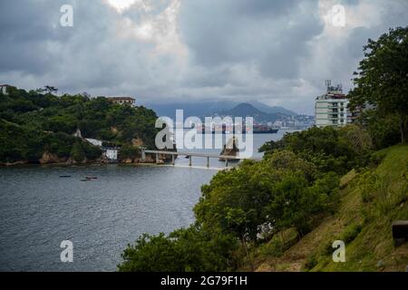 Der Blick von Ponte da Boa Viagem, Nitreói, Bundesstaat Rio de Janeiro, Brasilien. Grüne Ilha da Boa Viagem Privatinsel in Bucht, die über eine schmale Brücke und gelben Sand unter blauer Wolkenlandschaft mit dem Hauptland verbunden ist. Berge in der Ferne. Stockfoto