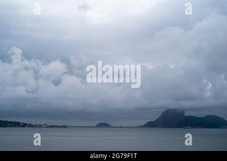 Der Blick von Ponte da Boa Viagem, Nitreói, Bundesstaat Rio de Janeiro, Brasilien. Grüne Ilha da Boa Viagem Privatinsel in Bucht, die über eine schmale Brücke und gelben Sand unter blauer Wolkenlandschaft mit dem Hauptland verbunden ist. Berge in der Ferne. Stockfoto