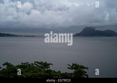 Der Blick von Ponte da Boa Viagem, Nitreói, Bundesstaat Rio de Janeiro, Brasilien. Grüne Ilha da Boa Viagem Privatinsel in Bucht, die über eine schmale Brücke und gelben Sand unter blauer Wolkenlandschaft mit dem Hauptland verbunden ist. Berge in der Ferne. Stockfoto