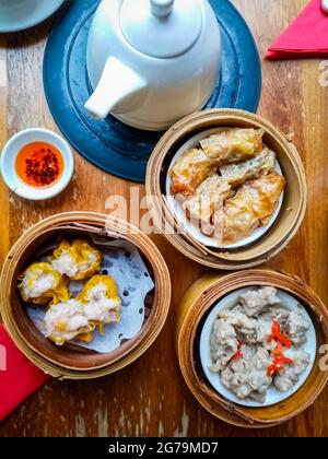 Blick von oben auf köstliche chinesische Dim-Sum-Gerichte in Bambusdampfer mit Teekocher und rotem Chilliöl. Stockfoto