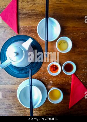 Blick von oben auf den Esstisch des chinesischen Restaurants mit Tee, Teekocher, weißen Schüsseln, Essstäbchen und rotem Chilliöl. Stockfoto