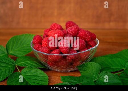 Reife große saftige Himbeeren in einem transparenten Teller auf einem Holztisch mit grünen Blättern. Stockfoto