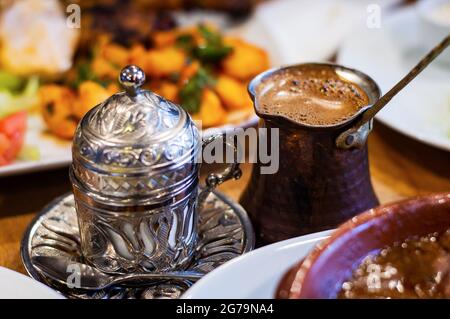 Türkischer Kaffee und traditioneller Metallbecher mit Prägedesign auf dem Tisch. Essen im Forground und Background verschwommen. Stockfoto