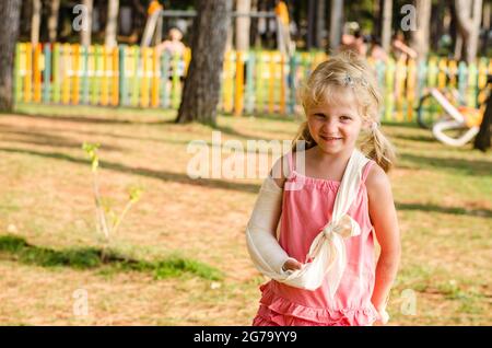 Schöne blonde Mädchen mit gebrochener Hand und Spielplatz im Hintergrund Stockfoto