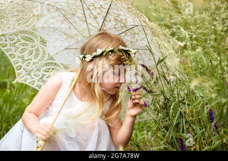 Mädchen mit langen Haaren hält Sonnenschirm riechende Blumen auf der Wiese Stockfoto