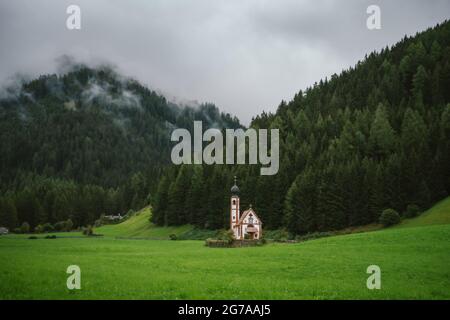 St. Johann Kirche in Ranui an bewölktem bewölktem, bewölktem Tag, Dolomiten-Alpen, Vilnosstal, Südtirol, Italien. Nebel über dem Kiefernwald Stockfoto