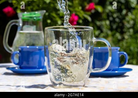 Wasser fließt in einen Becher mit einem Teebeutel. Ein Picknick im Sommergarten. Stockfoto