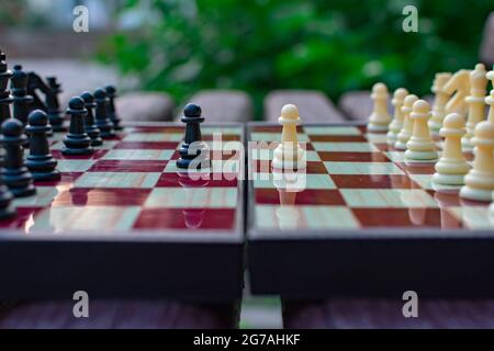 Schachfiguren werden auf einem Schachbrett auf einem unscharfen Hintergrund der Natur platziert. Die Bauern sind schwarz und weiß einander gegenüber. Unscharfer Fokus. Unschärfe-Effekte Stockfoto