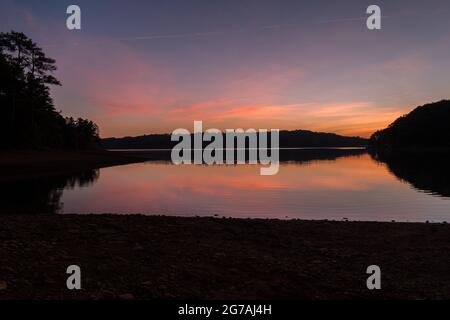 Sonnenaufgang hinter den Bäumen über dem See mit einer Spiegelung im bunten Himmel an einem frühen Morgen im Herbst Stockfoto