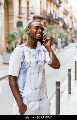 Vertikale Übernahme eines fröhlichen Afro-Mannes, der lächelt und telefoniert. Er kleidet sich legere Sommerkleidung an Stockfoto