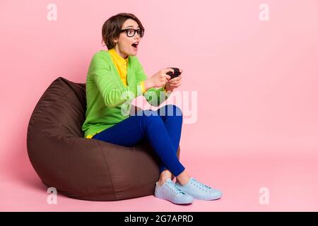 In voller Größe Foto von Brünette bob haarige Schulmädchen sitzen Stuhl spielen Konsole Spiel tragen Schuhe auf pastellrosa Hintergrund isoliert Stockfoto