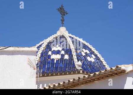 Typisch mediterrane blau-weiße Kuppel mit verziertem schwarzen schmiedeeisernen Kreuz unter klarem blauen Himmel. In touristischem Küstenort Calpe an der Costa Blanco. Stockfoto