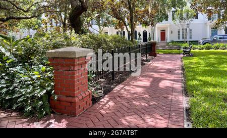 Ein Pfad aus rotem Backstein entlang eines Parks mit Bäumen, Metallzaun und Holzbänken im Stadtteil „The Cemebraging“ in Orlando, Florida. Stockfoto