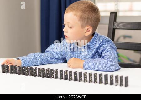 Der sechsjährige niedliche Junge spielt zu Hause auf einem weißen Holztisch Dominos. Selektiver Fokus. Nahaufnahme Stockfoto