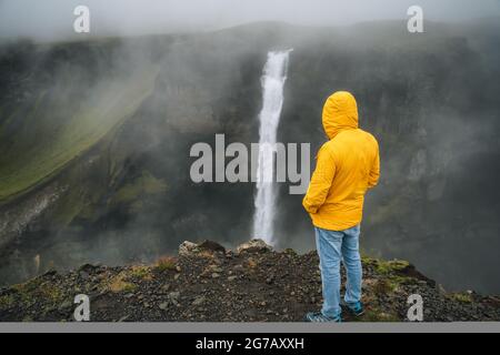 Ein Mann in gelber Jacke genießt den Haifoss Wasserfall bei regnerischem, bewölktem Wetter. Hochland von Island Stockfoto