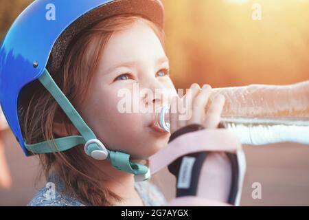 Ein kleines Mädchen in einem schützenden Rollschuh trinkt Wasser aus einer klaren Flasche. Hochwertige Fotos Stockfoto
