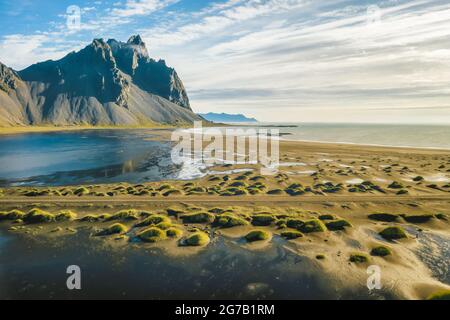 Blick von der fliegenden Drohne. Atemberaubende Szene von Stokksnes Cape mit Vestrahorn im Hintergrund. Isländische Panoramasicht auf schwarze Sanddünen bei Flut mit gelbem Gras Stockfoto