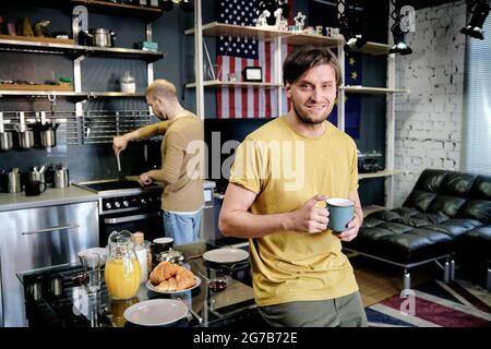 Schöner attraktiver lächelnder junger Mann trinkt Tasse Kaffee am Morgen, als sein Freund das Frühstück im Hintergrund kocht Stockfoto