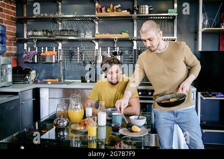 Junger Mann, der köstliche, flauschige Hüttenkäse-Pfannkuchen serviert, die er für seinen Mann gekocht hat Stockfoto