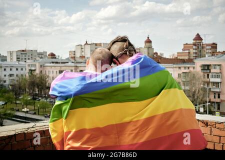 Rückansicht von zwei schwulen Männern, eingewickelt in Regenbogenfahne, die auf dem Dach stehen und die Stadt betrachten Stockfoto