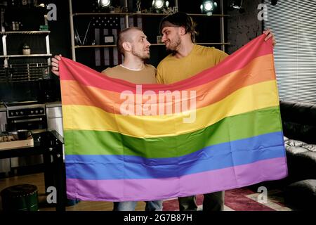 Junge liebevolle homosexuelle Männer halten Regenbogenfahne und schauen sich gegenseitig an, wenn sie in der Wohnung stehen Stockfoto
