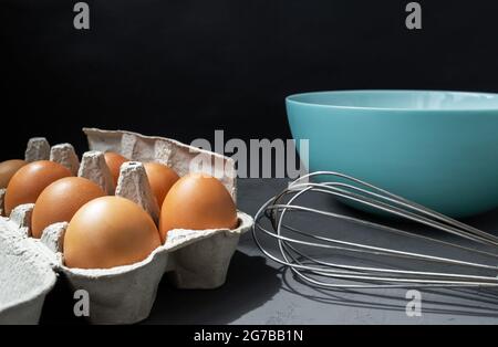 Frische Eier in Pappeiablage mit Schneebesen und Teller auf dunklem Grund Stockfoto