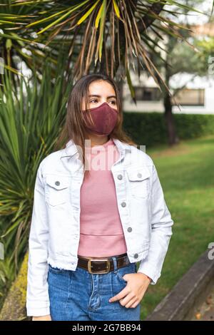 Vertikales Porträt eines Teenagers mit burgunderfarbener Maske, die Jean und eine rosa Bluse trägt und in einem Park im Freien nachdenklich aussieht Stockfoto