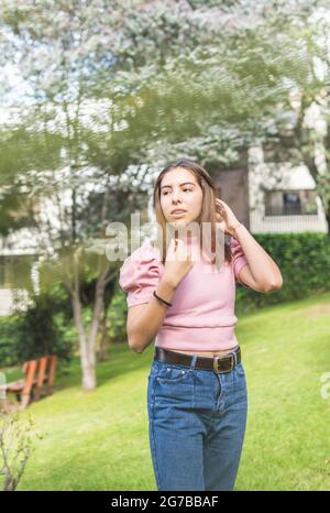 Outdoor-Porträt eines Latina-Teenagers mit Zahnspangen, die ihr gerades Haar mit ihren Händen in rosa Bluse und Jeans anpassen Stockfoto