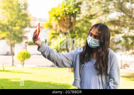 Brünette latinx Teenager-Mädchen mit OP-Maske, die ein Selfie mit ihrem Handy in einem Park Stockfoto