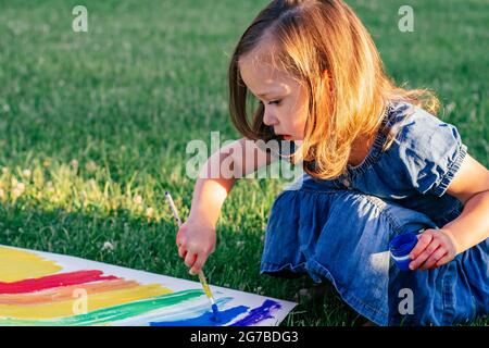 Kleines Mädchen 2-4 Jahre alt malt Regenbogen und Sonne auf ein großes Blatt Papier sitzen auf grünem Rasen in Sonnenlicht Stockfoto