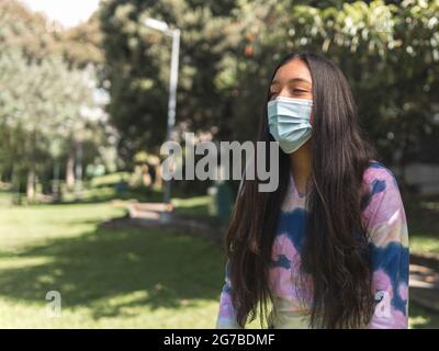 Teenagermädchen mit langen schwarzen Haaren und brauner Haut, die eine Maske trägt, lacht in einem Park mit Bäumen Stockfoto