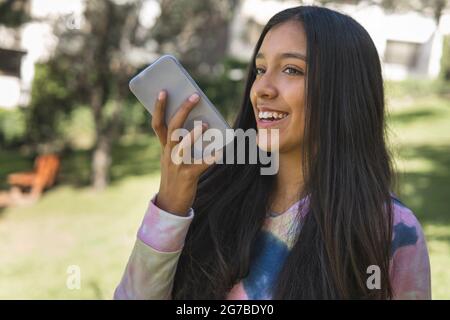 Fröhliches, lateinisches Teenager-Mädchen, das mit ihrem Handy eine Sprachnachricht sendet Stockfoto