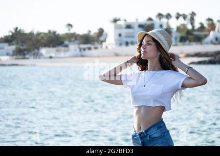 Glückliche attraktive hispanische junge Frau, die am Meer steht und einen Hut und Sommerkleidung trägt Stockfoto