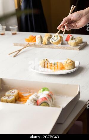 Hand einer Person, die mit Essstäbchen Sushi-Rolle in einem Pappkarton und Teller auf einem Tisch in einem Restaurant organisiert, Details der Essensstruktur, Studio Stockfoto