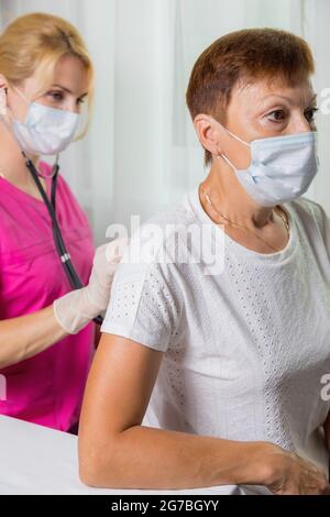 Eine Ärztin in einer Maske lauscht mit einem Stethoskop auf den Rücken einer erwachsenen Frau. Eine Frau in der Arztpraxis beschwert sich über Husten Stockfoto