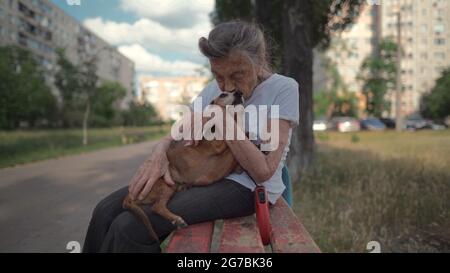 Ältere 90-jährige Frau mit grauem Haar und tiefen Falten sitzt im Freien in einer betreuten Wohnanlage auf der Bank mit einem kleinen Dackel-Hund. Alte weibliche Umarmungen Stockfoto