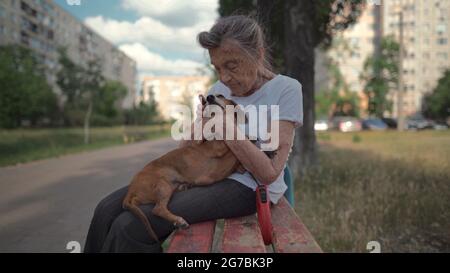 Ältere 90-jährige Frau mit grauem Haar und tiefen Falten sitzt im Freien in einer betreuten Wohnanlage auf der Bank mit einem kleinen Dackel-Hund. Alte weibliche Umarmungen Stockfoto