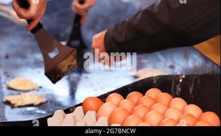 Ein Tablett oder Karton mit Eiern im Vordergrund mit einem Kochfeld bbq im Hintergrund. Hände und Kochutensilien drehen Spiegeleier. Spendenaktion für die Gemeinschaft Stockfoto