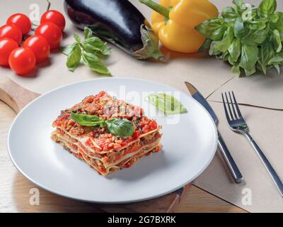 Lasagne auf weißem Teller. Basilikum, Tomaten, Zucchini und Pfeffer im Hintergrund. Speisekarte mit italienischer Küche. Stockfoto