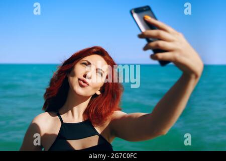 Das rothaarige Mädchen lässt ihre Lippen entweichen und macht Selfie auf der Smartphone-Kamera. Hübsche Frau im schwarzen Badeanzug fotografiert sich vor dem Hintergrund von Meer und Himmel. Sommer sonniger Urlaub. Stockfoto