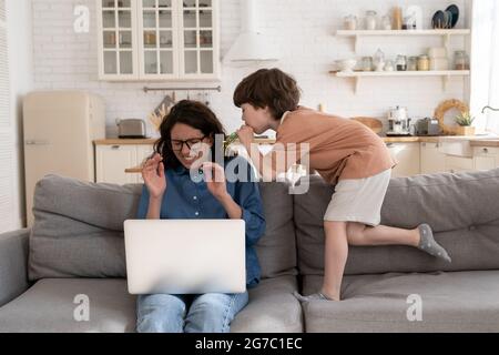 Erschöpfte alleinerziehende Mutter arbeitet von zu Hause aus mit einem hartnäckigen kleinen Vorschulkinder-Sohn vom Laptop Stockfoto