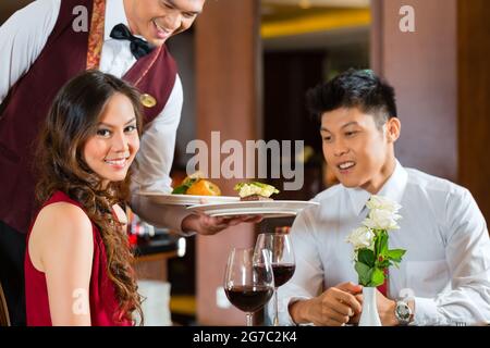 Asiatische chinesische Paar - Mann und Frau - oder Liebhaber mit einem Datum oder einem romantischen Abendessen in einem ausgefallenen Restaurant während der Kellner ist das Essen Stockfoto