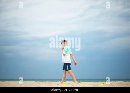 Boy Training an einem Sandstrand. Tennisbälle werden an der Küste verteilt. Ruhe zwischen den Übungen. Stockfoto